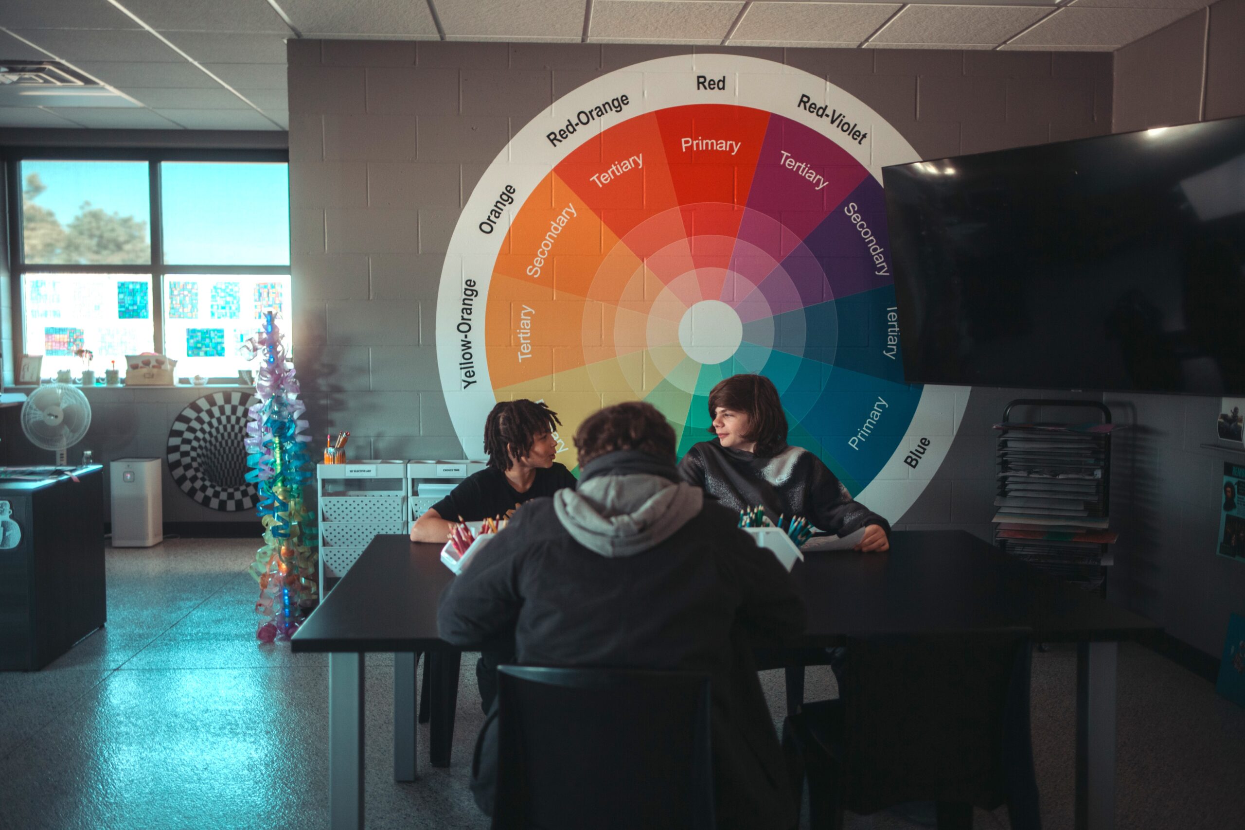 Three boys sit at a table in front of a large color wheel talking and doing art together.